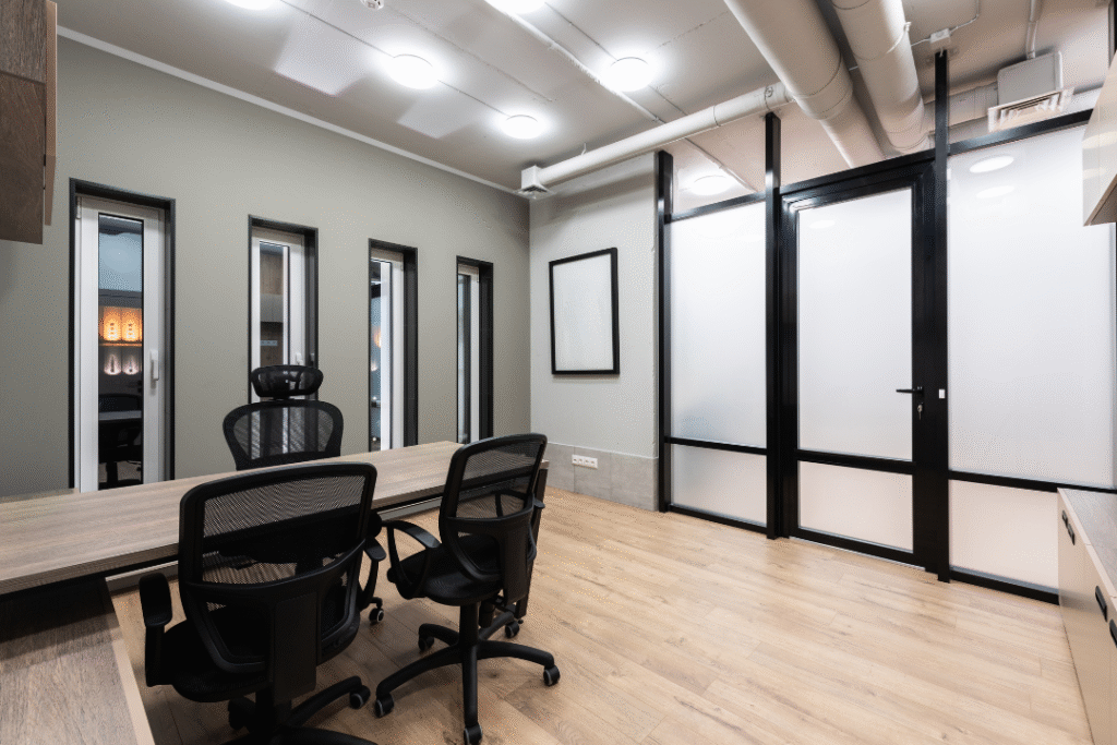 A modern office with wooden floors, a long desk, three black office chairs, gray walls, and large black-framed glass partitions separating spaces. The room is well-lit with ceiling lights.