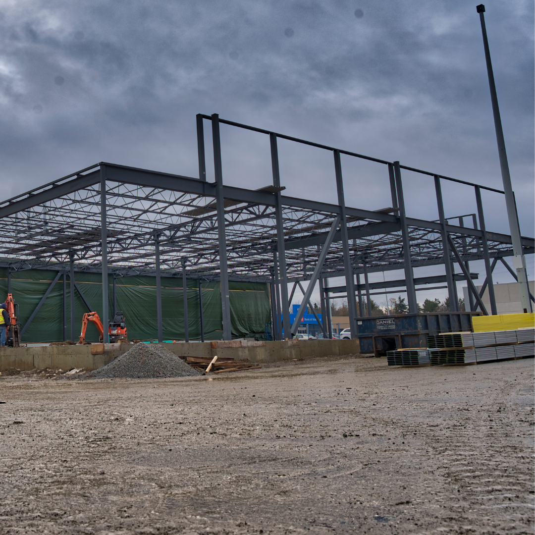 A steel framework of a building under construction stands on a muddy site with construction materials and equipment nearby under a cloudy sky.