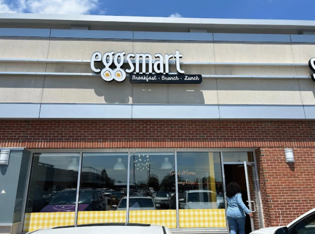 A person walks toward the entrance of an EggsMart restaurant, which has a sign reading “eggsmart: Breakfast • Brunch • Lunch” above its glass windows and brick facade. Several cars are parked in front.