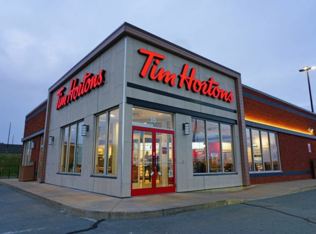 A Tim Hortons restaurant with large windows and bright red signage is seen at dusk, with lights on inside and an empty parking lot in front.