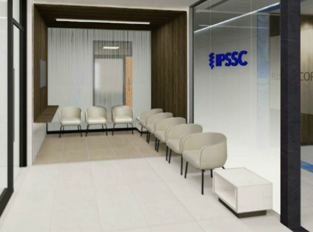 A modern waiting room with beige chairs lining two walls, a small white table, wood and glass accents, and IPSSC displayed in blue on a glass partition. The space is clean and well-lit.