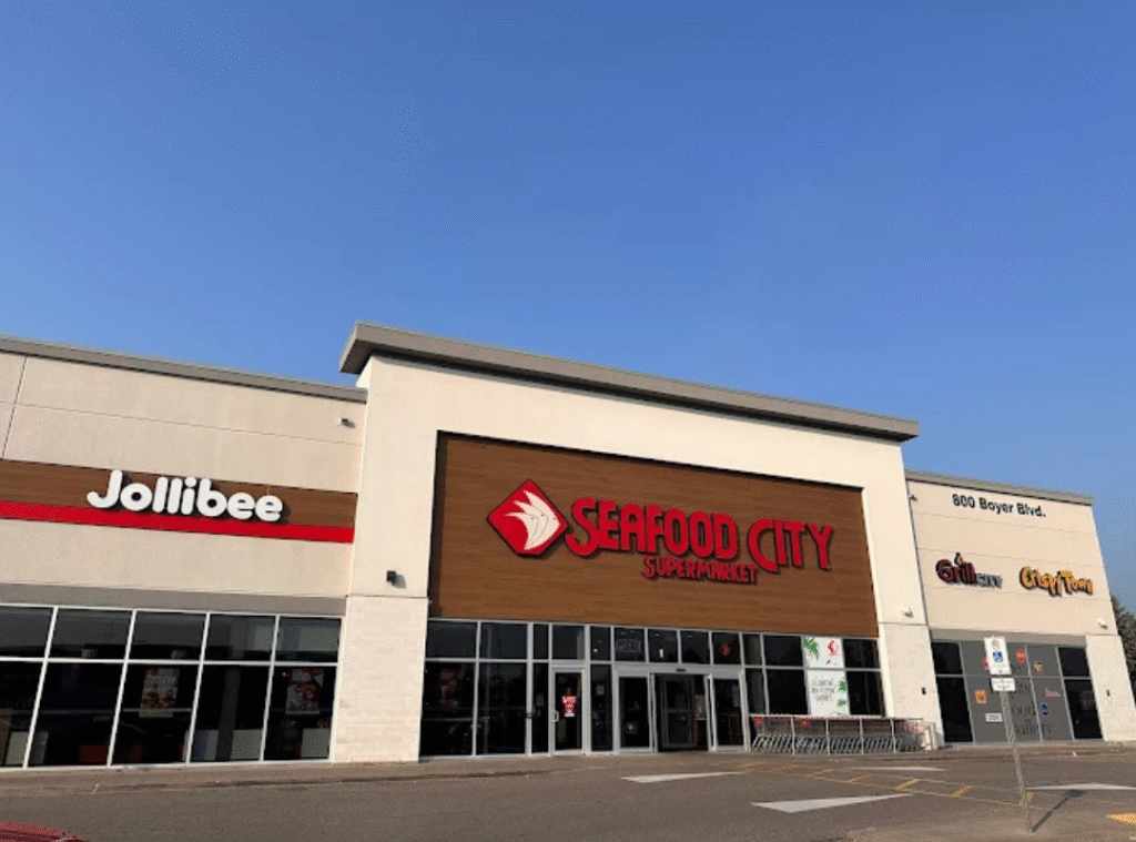 A shopping plaza with Jollibee on the left, Seafood City Supermarket in the center, and additional stores on the right under a clear blue sky. Shopping carts are lined up near the entrance.