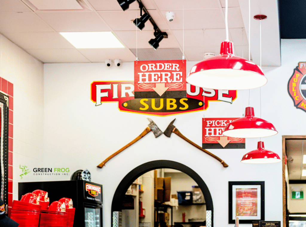 Interior of a Firehouse Subs restaurant showing red hanging lights, a large Order Here sign above an arched counter, fire axes on the wall, and the Green Frog Construction Inc. logo on the left.