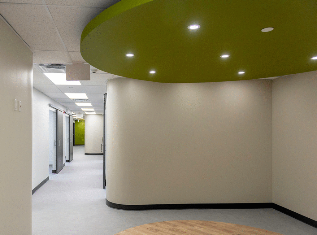 A modern office hallway with cream-colored walls, gray flooring, a curved green ceiling with recessed lights, and several doorways along the left side.