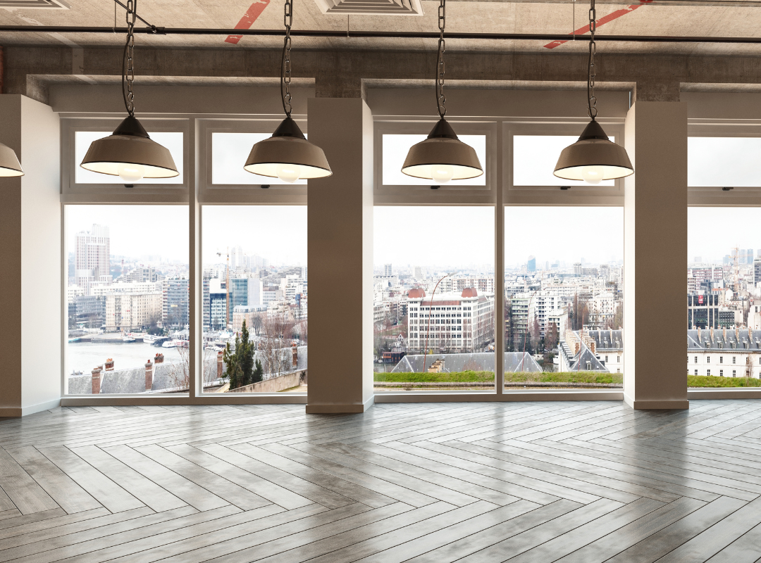 Spacious, empty room with large windows, pendant lights, and a view of a city skyline and river. The room has wooden herringbone flooring and an industrial-style ceiling.